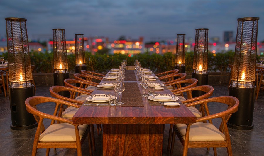 Rooftop restaurant terrace at dusk with a long teak dining table, chairs and glowing patio heaters above city lights