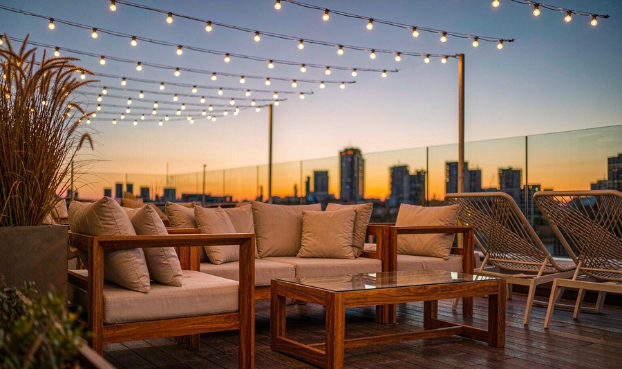 Rooftop bar terrace at golden hour with teak sofa set, rope lounge chairs and string lights above a city skyline
