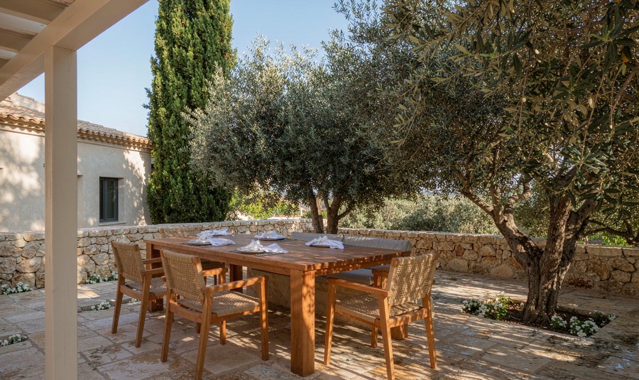 Villa garden terrace with a long teak dining table and woven-back chairs in dappled afternoon light